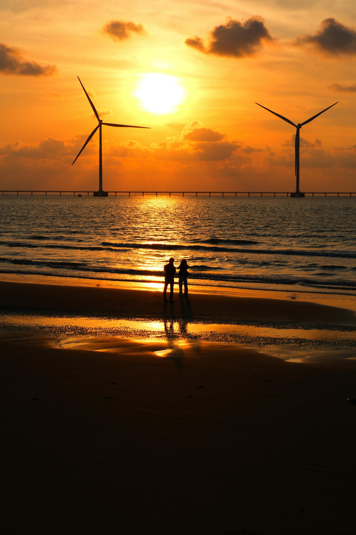 Silhouetted couple on a beach with wind turbines against a stunning sunset backdrop.